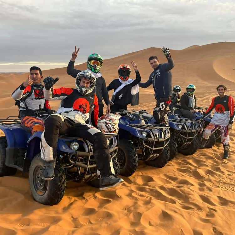Merzouga Quad Bike group of riders in colorful gear on blue quad bikes posing on golden sand dunes under cloudy sky