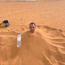 Man partially buried in reddish desert sand with only his head visible, demonstrating Merzouga Sand Baths traditional therapy.