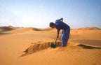 Man in blue clothing digging warm golden sand in desert dunes for Merzouga Sand Baths traditional therapy experience
