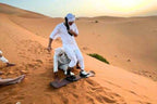 Sandboard with black straps on golden sand dunes with two men in white clothing preparing to sandboard, showcasing Merzouga Sandboarding.