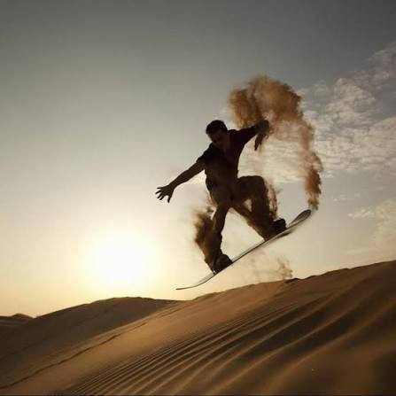 Sandboarder in mid-air kicking up golden sand dunes at sunset during Merzouga Sandboarding in the Sahara Desert