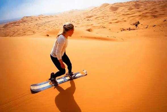 Merzouga Sandboarding showing a woman in white top and black pants gliding down golden sand dunes on a blue and white sandboard.