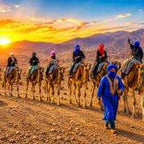 Merzouga sunrise camel trekking with a group of riders on camels led by a guide in blue robe across golden desert dunes.