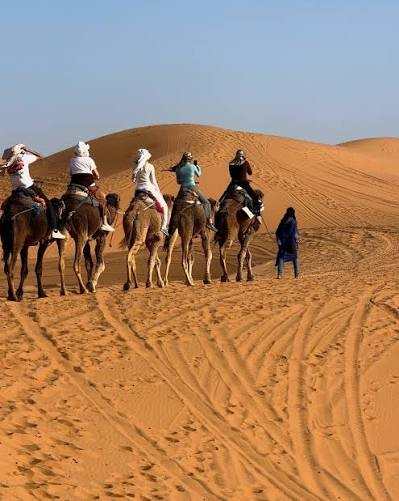 Camel trekking adventure with riders and a guide in blue robe across golden sand dunes during Merzouga sunrise in the Sahara Desert