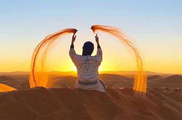 Person in white clothing throwing sand upwards in golden desert dunes at Merzouga sunrise with clear sky background