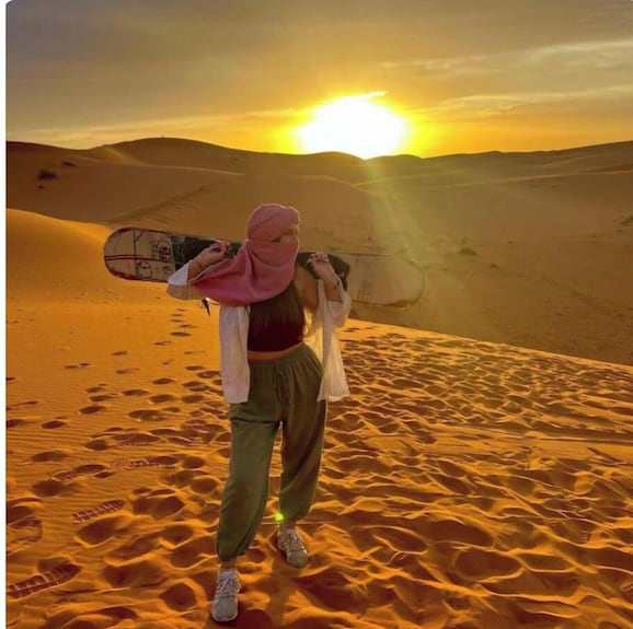 Merzouga Sunset Camel Trek featuring a person in casual attire carrying a sandboard at golden desert dunes during sunset
