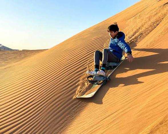 Young person in blue jacket sandboarding down golden sand dune at sunset during Merzouga Sunset Camel Trek adventure