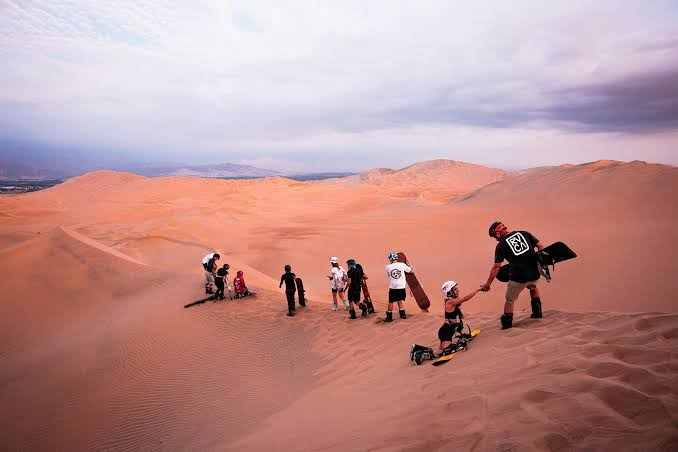 Merzouga Sunset Camel Trek showing group of people sandboarding and walking on vast golden sand dunes under cloudy sky