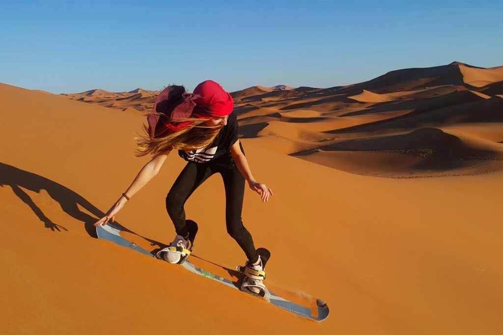 Woman sandboarding on golden sand dunes under clear blue sky during Merzouga Sunset Camel Trek adventure