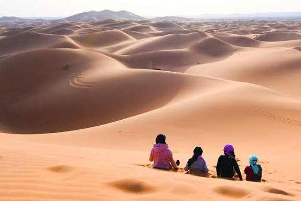Expansive golden sand dunes with four people sitting on the sand wearing colorful clothing during Merzouga Sunset Camel Trekking.