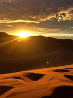 Sunset over golden sand dunes with dramatic cloud patterns highlighting the Merzouga Sunset Camel Trekking landscape.