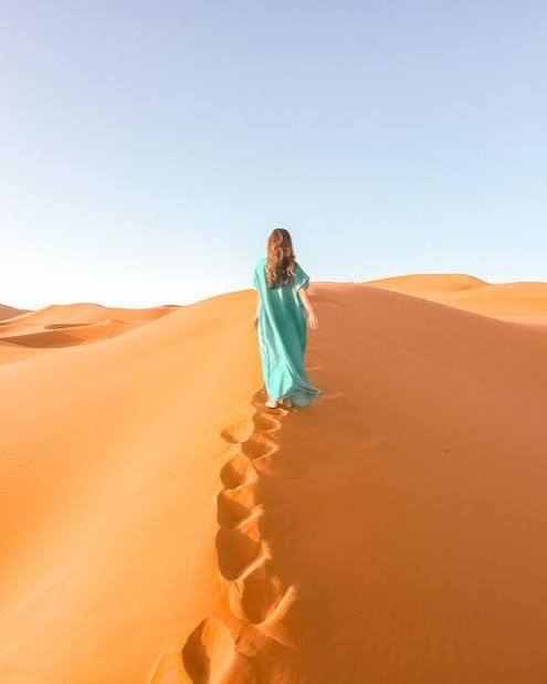 Woman in a turquoise dress walking up golden sand dunes under clear sky during Merzouga Sunset Camel Trekking adventure