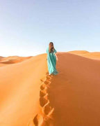 Woman in a turquoise dress walking up golden sand dunes under clear sky during Merzouga Sunset Camel Trekking adventure