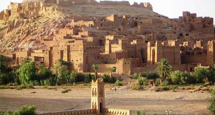 Ancient desert village with brown earthen buildings and palm trees in a rocky landscape under sunlight, representing Merzouga Taxi Rent travel area