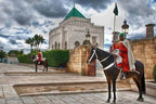 Historic Moroccan palace with traditional architecture guarded by two soldiers on horses in red and white ceremonial uniforms under a cloudy sky, illustrating Merzouga Taxi Rent cultural journey.