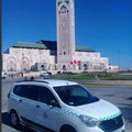 White compact taxi car with green checker pattern on sides and hood parked near a large mosque with a tall minaret, in Morocco, representing Merzouga Taxi Rent.