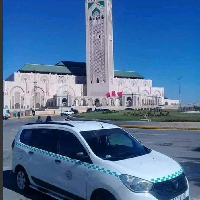 White compact taxi car with green checker pattern on sides and hood parked near a large mosque with a tall minaret, in Morocco, representing Merzouga Taxi Rent.