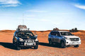 Two off-road vehicles, one black with roof luggage and the other white, parked on reddish desert sand under a clear blue sky, ideal for Day Tour from Fes to Merzouga.