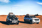 Two off-road vehicles, one black with roof luggage and the other white, parked on reddish desert sand under a clear blue sky, ideal for Day Tour from Fes to Merzouga.