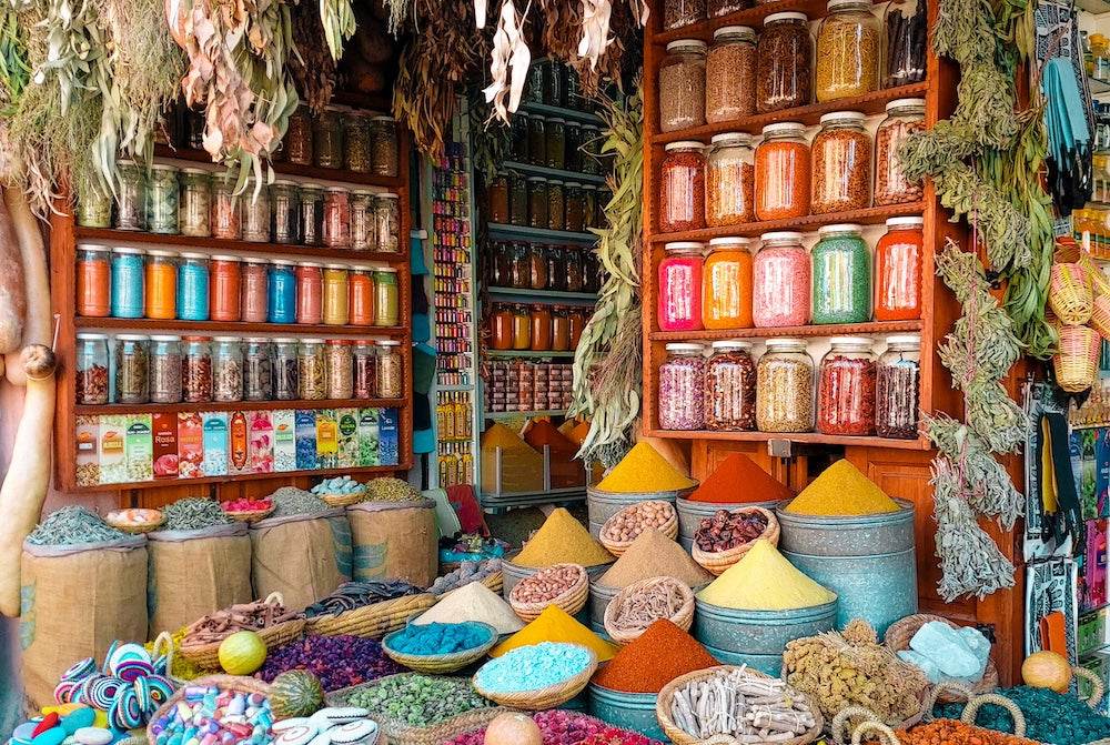 Spice shop with colorful powdered spices in conical piles, jars of spices on shelves, and hanging dried herbs