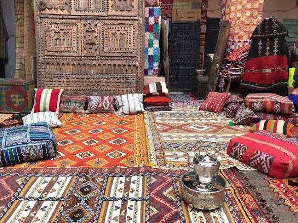 Traditional Moroccan rugs and cushions in various vibrant colors and geometric patterns with a metal tea set on a tray, evoking Merzouga to Fes cultural style