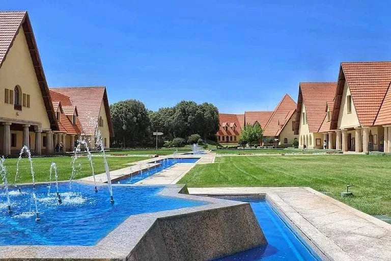 Traditional beige buildings with red tiled roofs surrounding a blue water fountain and manicured green lawn under clear blue sky in Merzouga to Fes tour