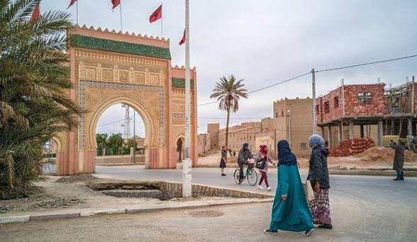 Traditional Moroccan city entrance arch with green tiled roof and pink walls, flags flying, people walking nearby under cloudy sky on Merzouga to Fes tour