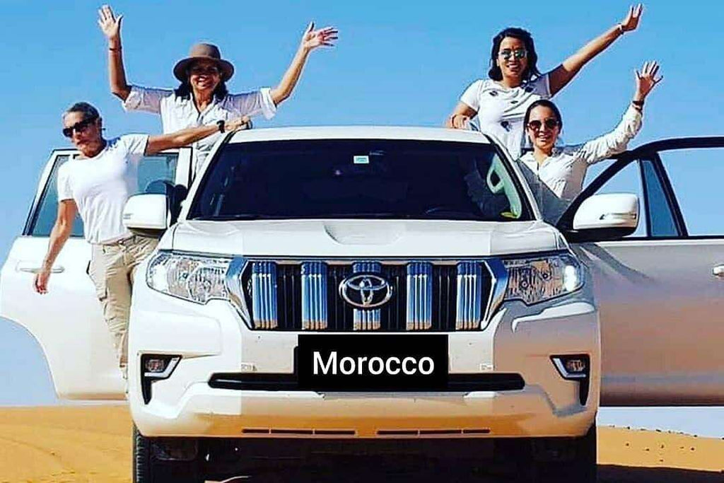 White Toyota SUV with four smiling women posing with arms raised standing by open doors in desert sand, representing Merzouga to Fes tour