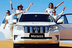 White Toyota SUV with four smiling women posing with arms raised standing by open doors in desert sand, representing Merzouga to Fes tour