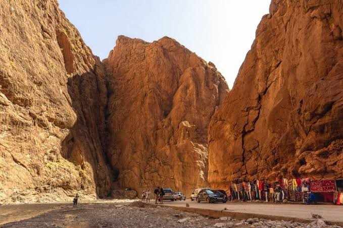 Todra Gorge with towering orange-brown rocky cliffs, a dry riverbed, cars parked by roadside and colorful textiles displayed on wall