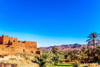 Ancient red mud-brick fortress beside a lush green palm oasis with mountain backdrop under a clear blue sky, showcasing Merzouga to Zagora desert landscape.