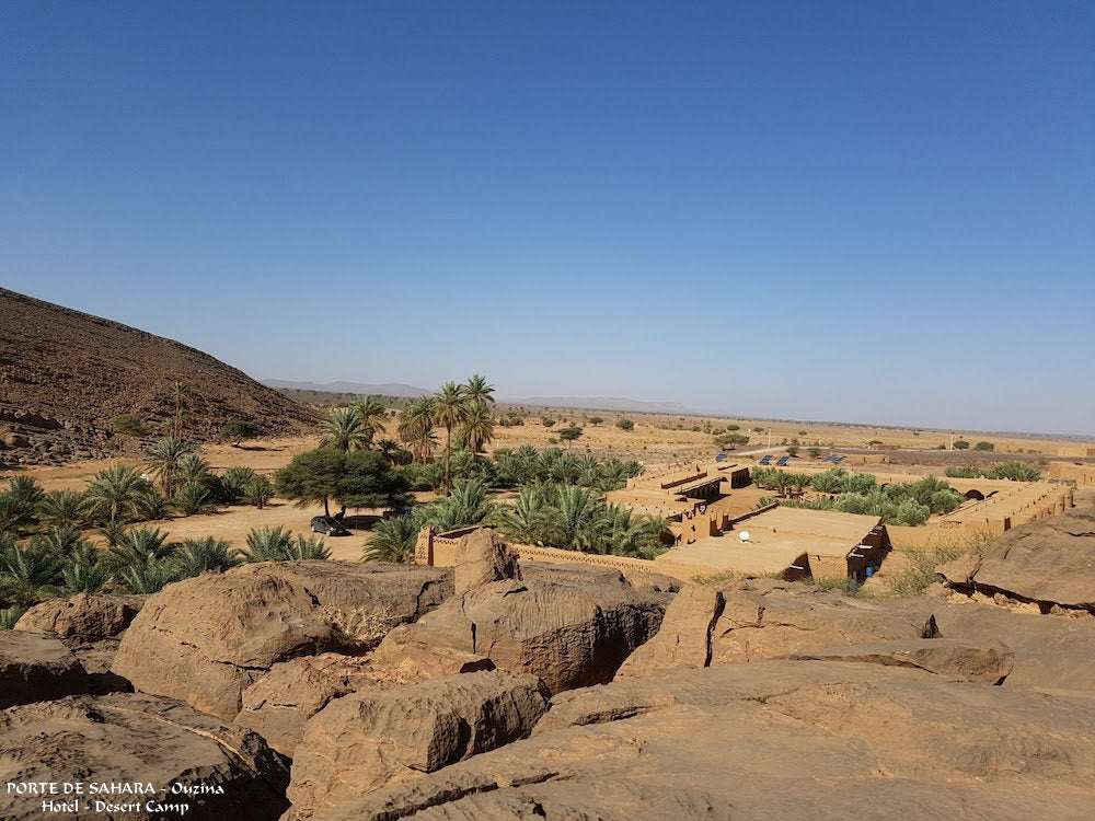 Desert landscape with rocky foreground, sandy terrain, green palm trees, and traditional mud-brick buildings showcasing Merzouga to Zagora scenery.