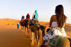 Camel caravan with riders in colorful clothing crossing golden sand dunes at sunset, showcasing a Merzouga Tour Guide experience.