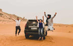 Three people jumping cheerfully beside a black SUV on orange sand dunes under a clear sky, embodying a lively Merzouga Tour Guide experience.