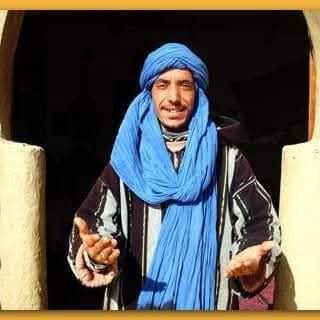 Merzouga Tour Guide wearing a blue turban and a black and white striped traditional Moroccan robe standing with open hands.