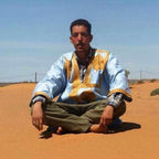Man wearing a light blue and yellow traditional robe sitting cross-legged on sand dunes, Merzouga Tour Guide.