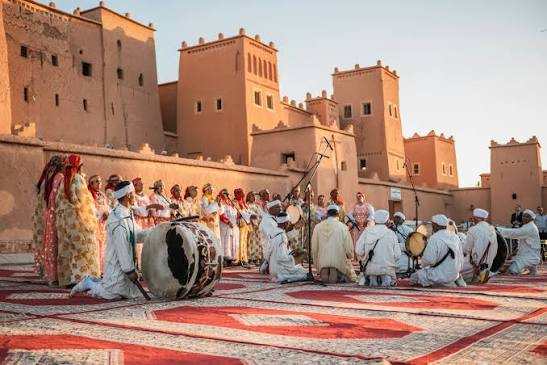 Traditional Moroccan musicians and singers dressed in white and colorful robes perform on patterned red and beige carpets in a desert village, reflecting cultural heritage with clay buildings in the background, highlighting a Merzouga Tour Guide experience.