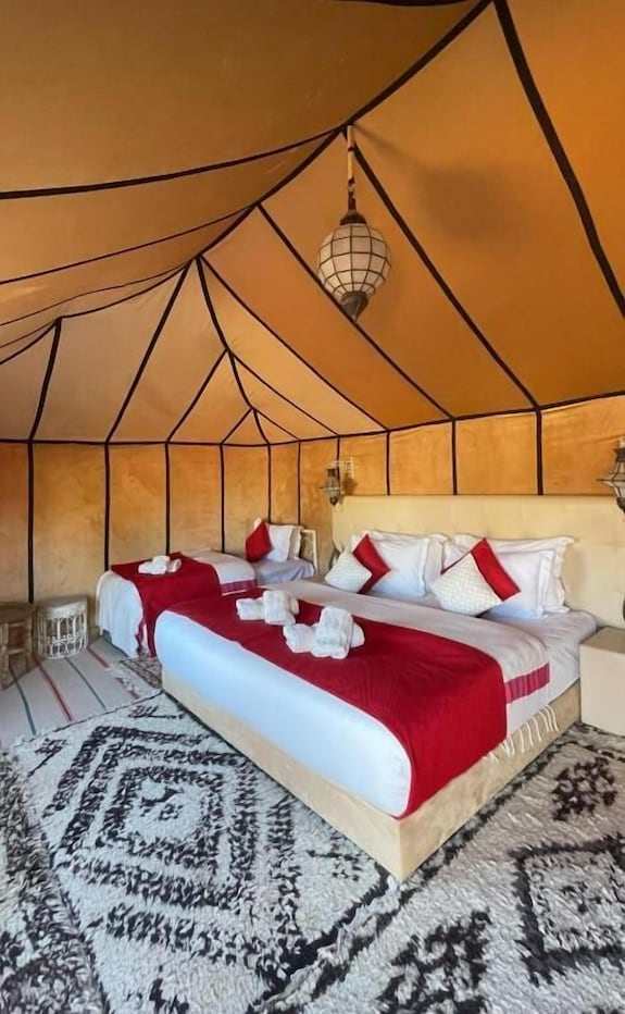 Merzouga VIP Luxury Camp bedroom with beige canvas tent, large bed and single bed, red accents, white linens, and patterned rug
