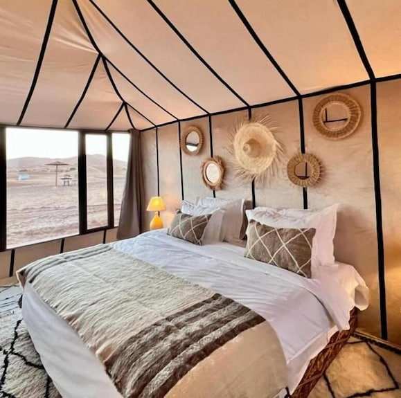 Merzouga VIP Luxury Camp interior featuring a beige and black striped tent with a large bed adorned with white and brown linens, decorative pillows, and woven wall ornaments