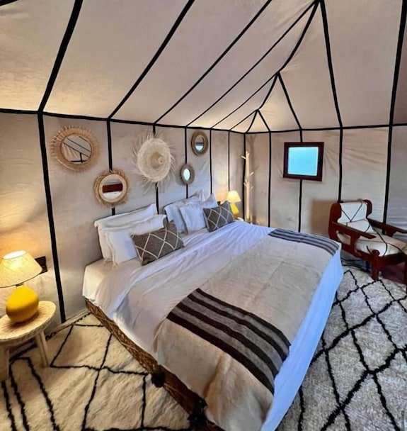 Merzouga VIP Luxury Camp spacious desert tent interior with large bed, white linens, black striped blanket, woven wall mirrors, and cozy lighting