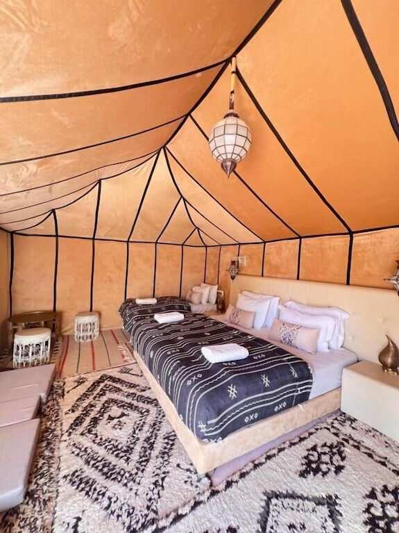 Merzouga VIP Luxury Camp spacious beige desert tent with black striped walls, twin beds covered in patterned black and white throws, and traditional lantern lighting