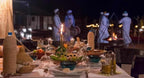 Merzouga VIP Luxury Camp evening dining setup with decorated table, glassware, woven bottles, salad plates, and performers in traditional attire
