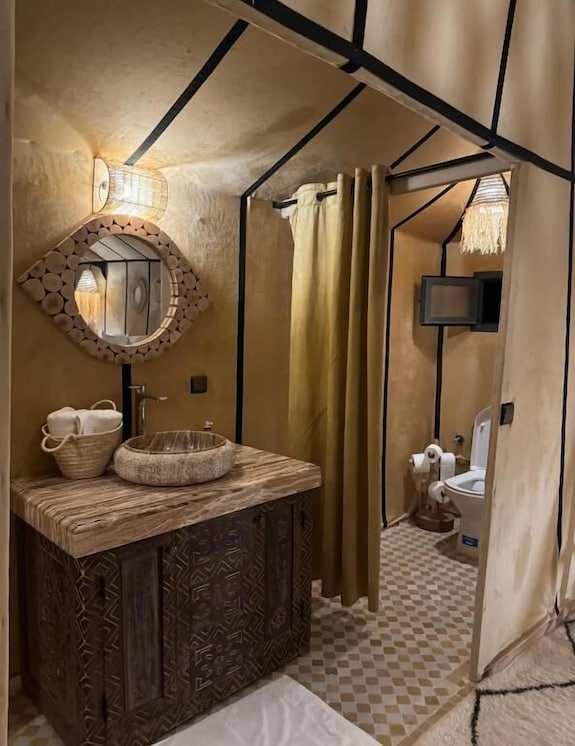 Merzouga VIP Luxury Camp rustic bathroom with carved wooden vanity, stone sink, round mirror, beige walls, and mustard shower curtain