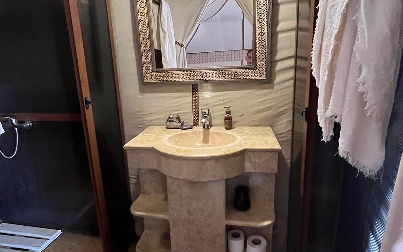 Bathroom sink with beige stone countertop and ornate framed mirror, towels hanging and toilet paper visible, Moonlight luxury Desert Camp