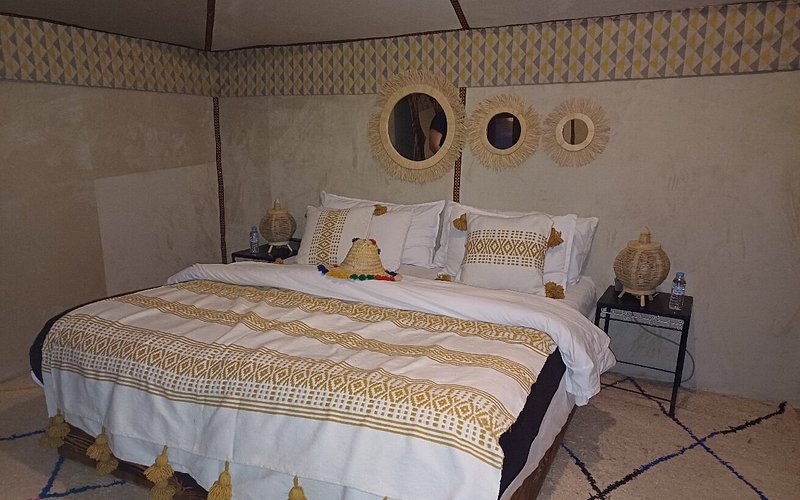 Luxury tent bedroom at Moonlight luxury Desert Camp featuring a large bed with white and brown patterned linens, decorative pillows, rustic woven mirrors, and bedside wicker lamps