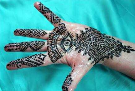 Hand with a detailed black Moroccan Henna Tattoo featuring geometric and floral patterns on a light skin tone against a turquoise background.