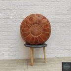 Moroccan pouf in brown leather with detailed stitched geometric patterns and a zipper closure, displayed on a small wooden stool