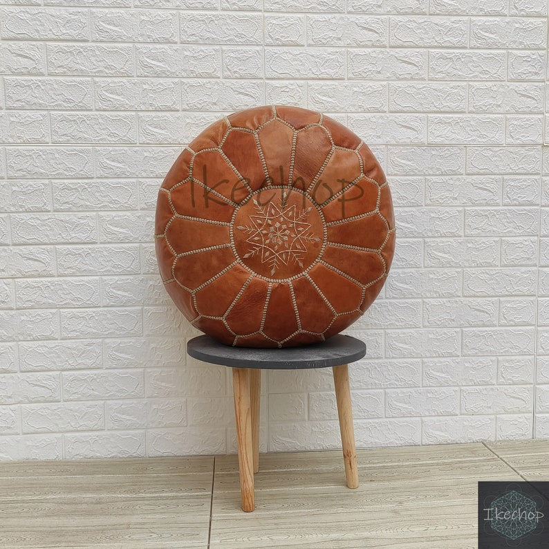 Moroccan pouf in brown leather with detailed stitched geometric patterns and a zipper closure, displayed on a small wooden stool