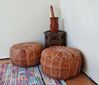 Moroccan pouf set in brown leather with white stitch detailing and zip closure, placed on a colorful woven rug