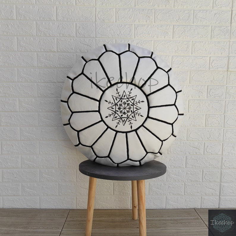 Round Moroccan pouf in white fabric with black embroidered geometric patterns and central star design, placed on a wooden stool
